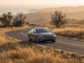 Black Tesla car driving on a winding road through a dry, grassy landscape at sunset.