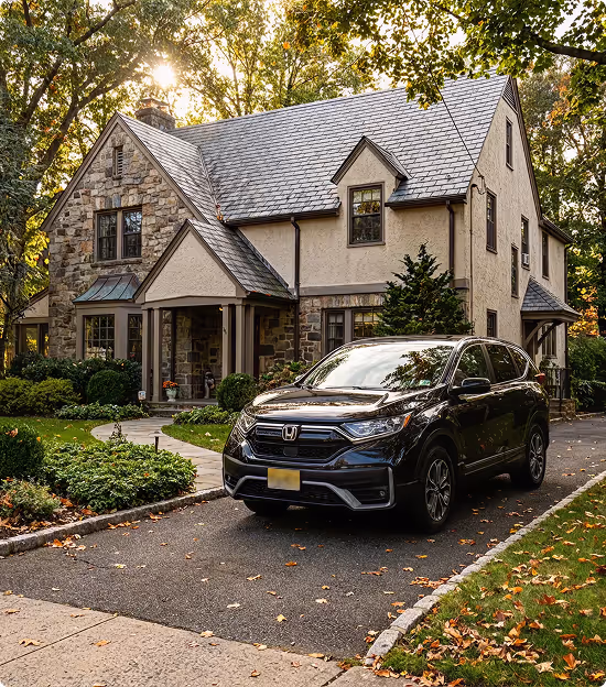 Black Honda SUV parked in the driveway of a two-story stone and stucco house surrounded by trees and greenery.