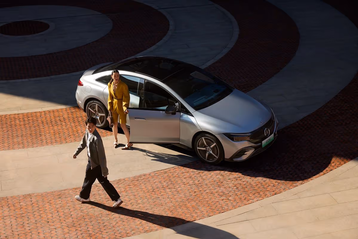 Woman in yellow suit standing next to silver Mercedes-Benz car with open door while a man walks nearby on brick and stone pavement.