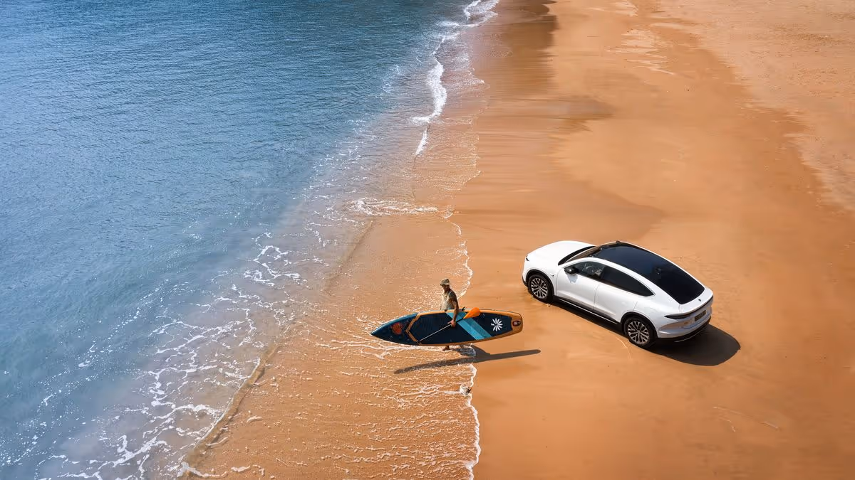 A person holding a paddleboard walking from a white car parked on a sandy beach towards the ocean.