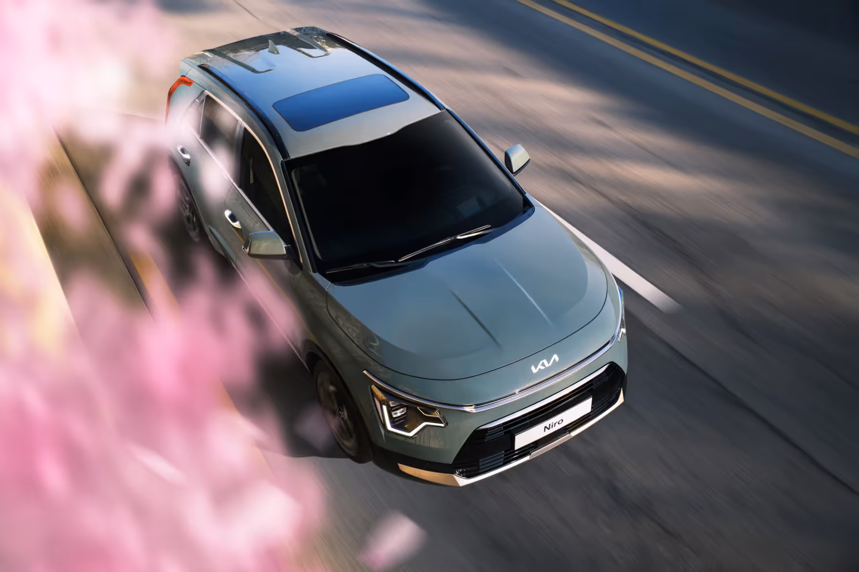 Silver Kia Niro SUV driving on road with pink blurred flowers in foreground.