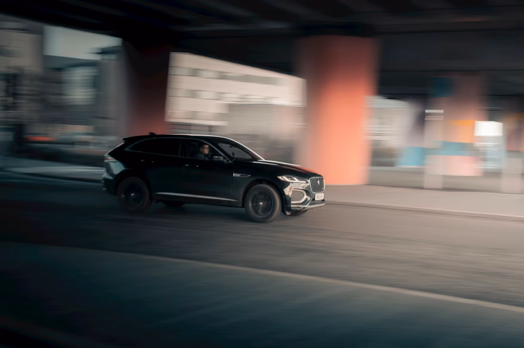 Black SUV driving fast under an overpass with blurred background cityscape.