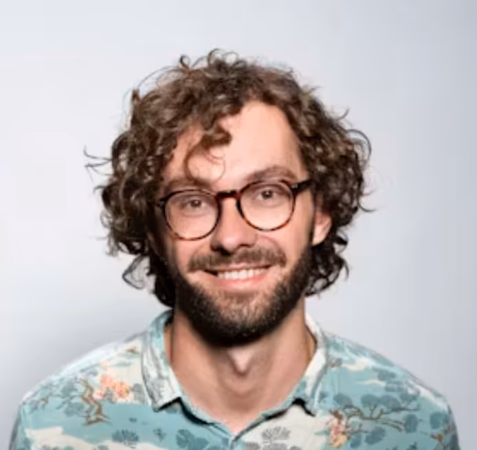 Smiling man with curly hair, beard, glasses, and floral shirt against a plain light background.