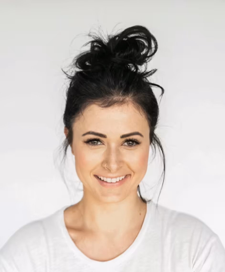 Smiling woman with dark hair in a messy bun wearing a white top against a white background.