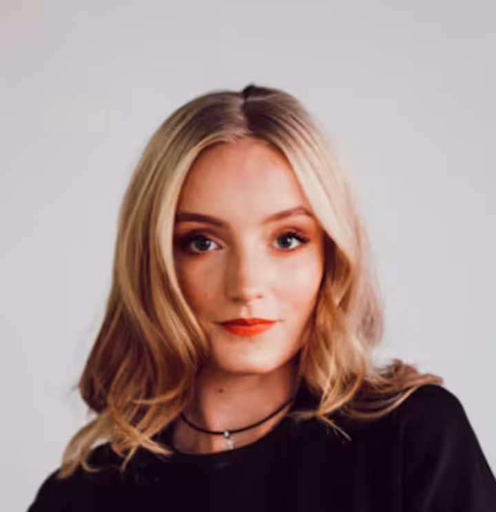 Portrait of a young woman with blonde hair, red lipstick, and black top against a light background.
