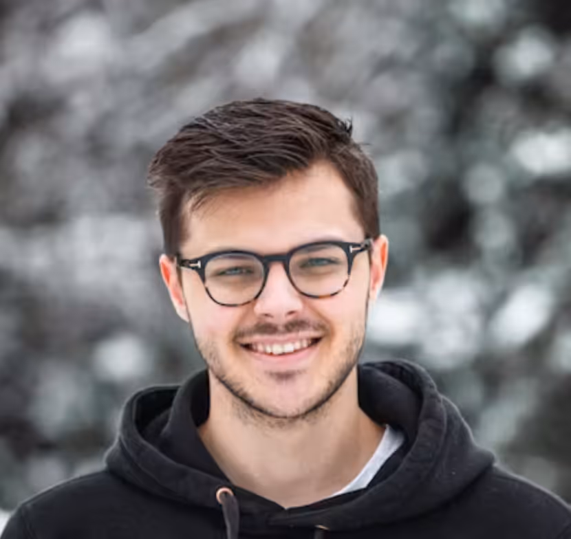 Smiling young man with glasses wearing a black hoodie with a blurred gray background.