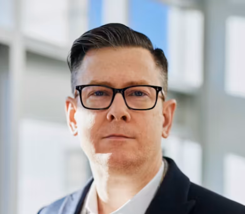 Man with dark hair and glasses wearing a dark jacket and white shirt in a bright indoor setting.