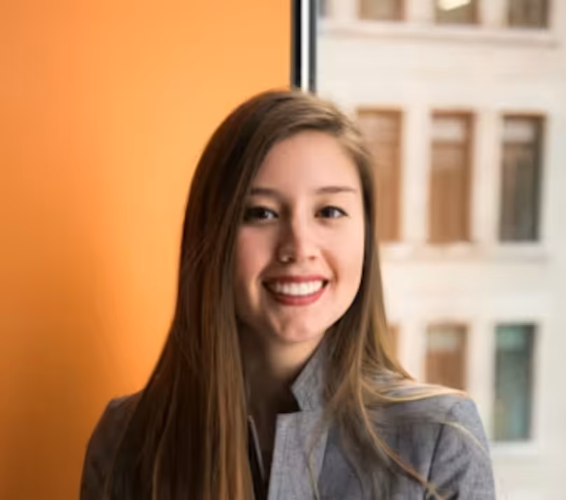 Young woman with long brown hair smiling, standing indoors with an orange wall and window behind her.