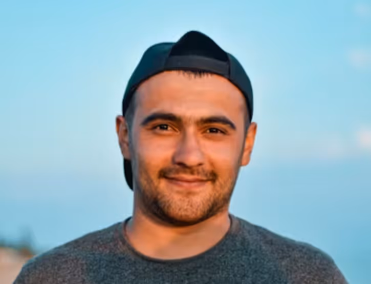 Smiling man wearing a backward black cap and gray shirt against a blue sky background.