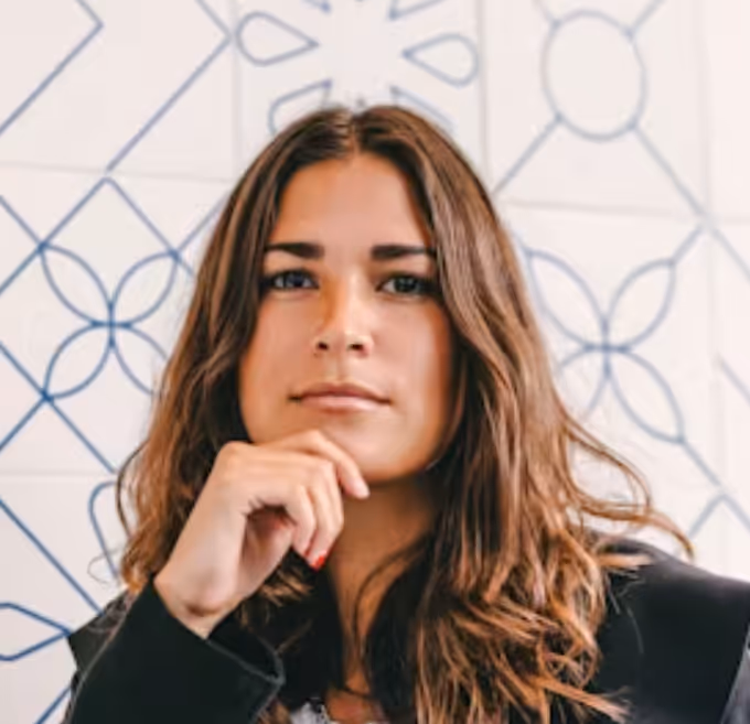 Woman with long brown hair resting her chin on her hand against a patterned white wall.