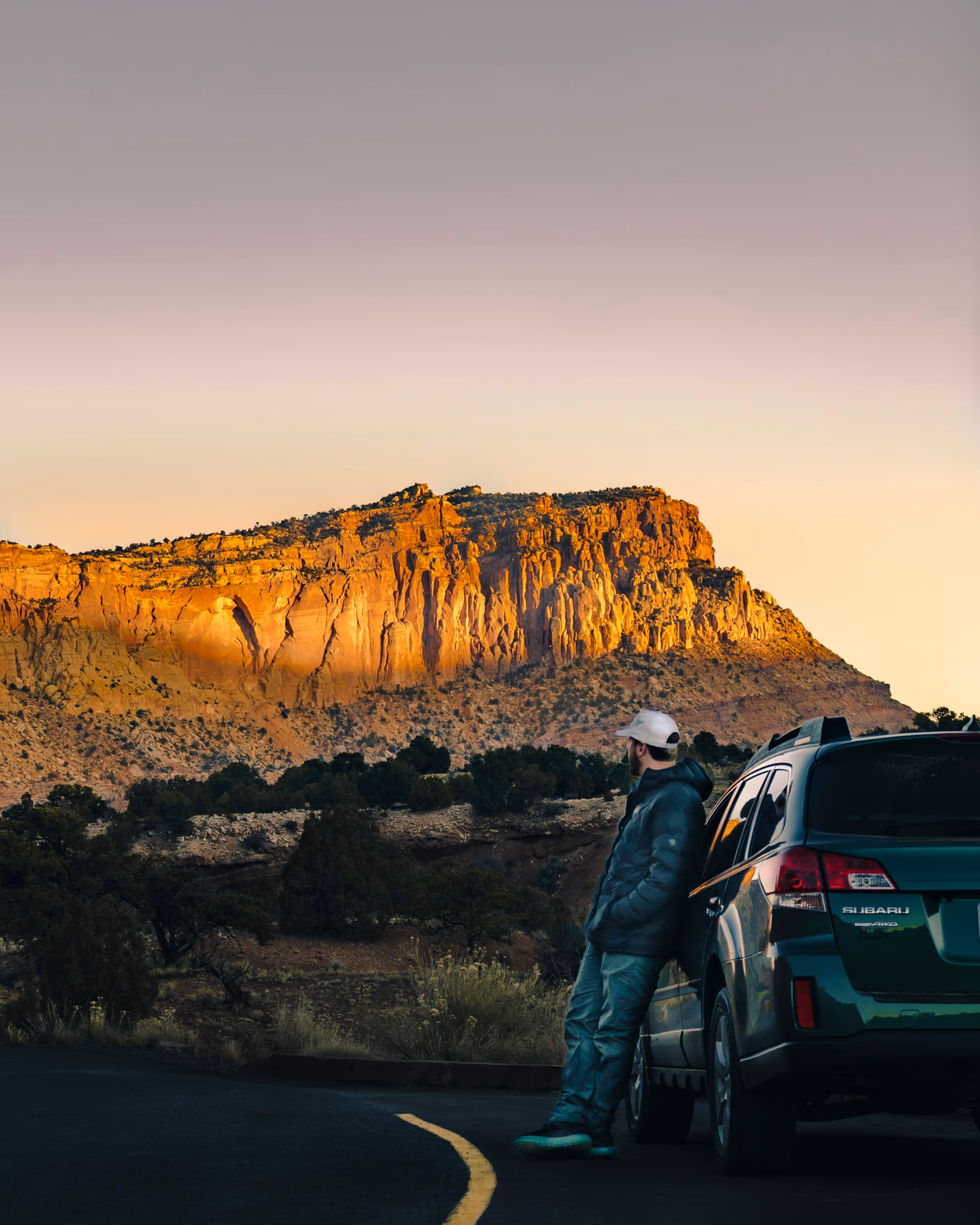Man leaning on a green Subaru parked beside a road, looking at sunlit rocky cliffs at sunset.