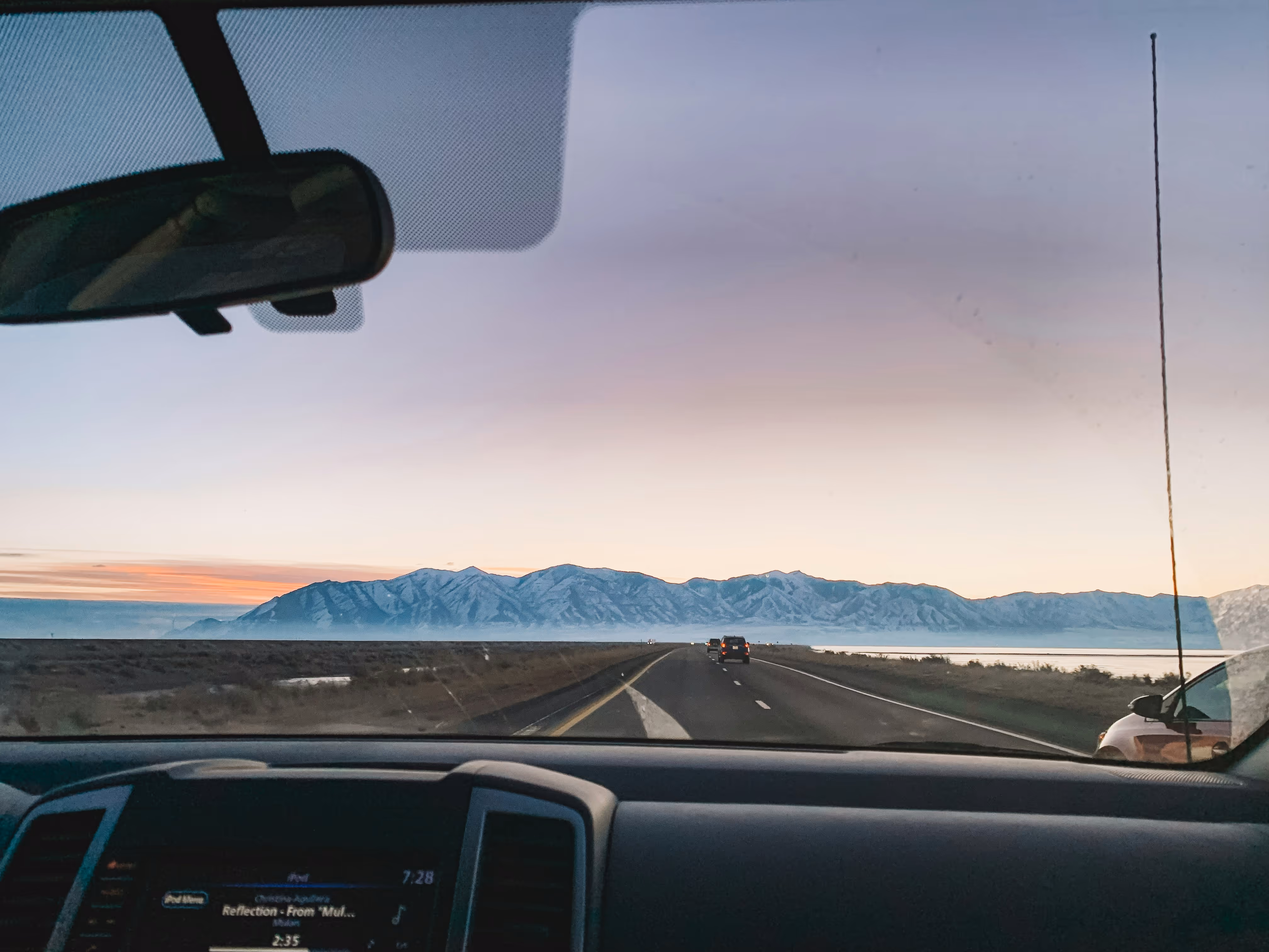 View through car windshield of highway leading to snow-capped mountains at sunset.