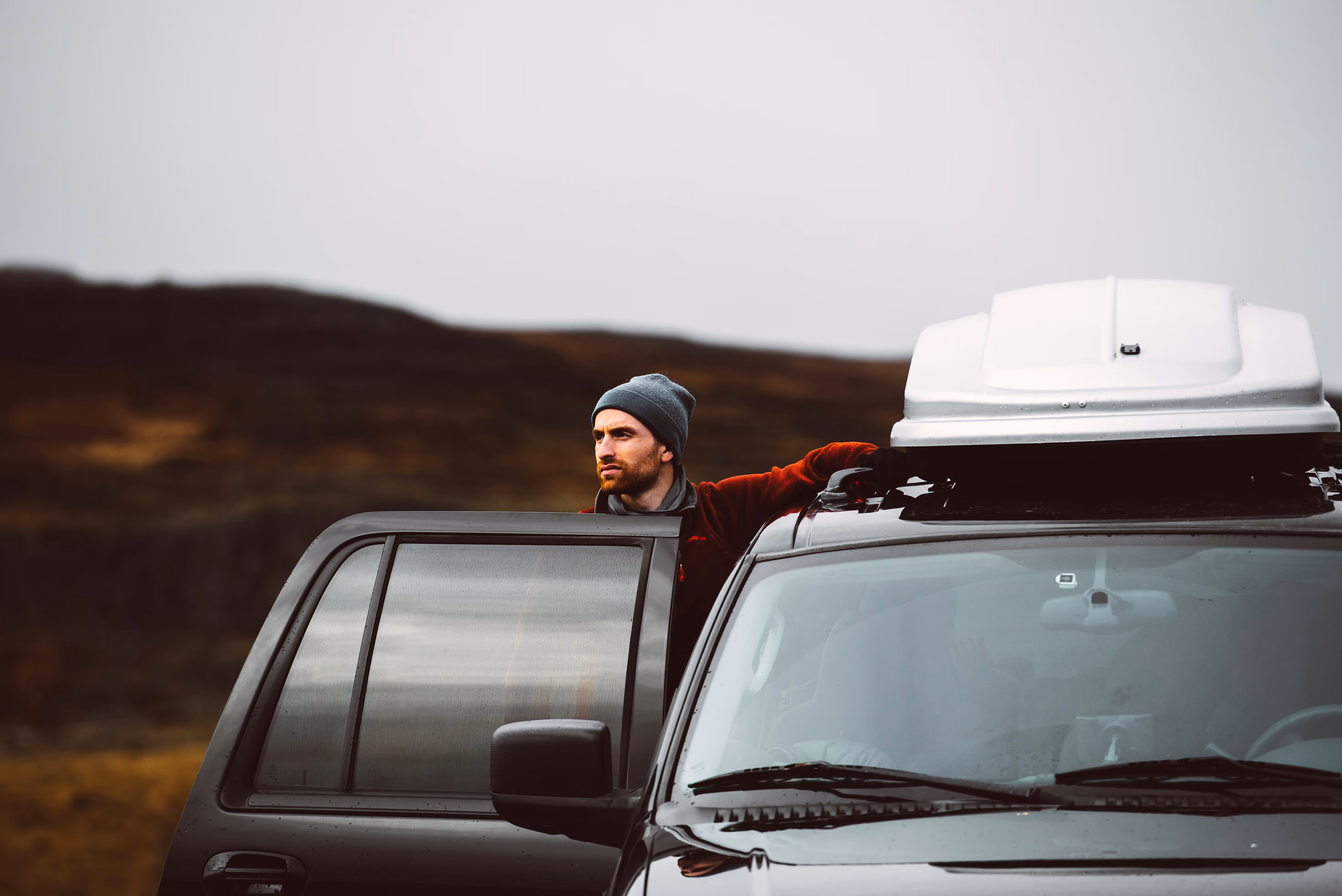 Man in a gray beanie standing outside black SUV with roof storage in a natural landscape.