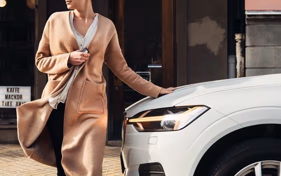 Person in a light brown cardigan standing next to a white car with modern headlights, touching the car's hood.