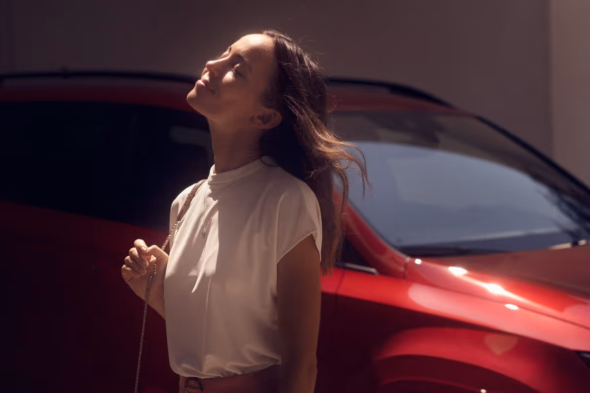 Woman standing with eyes closed, enjoying sunlight with a red car in the background.