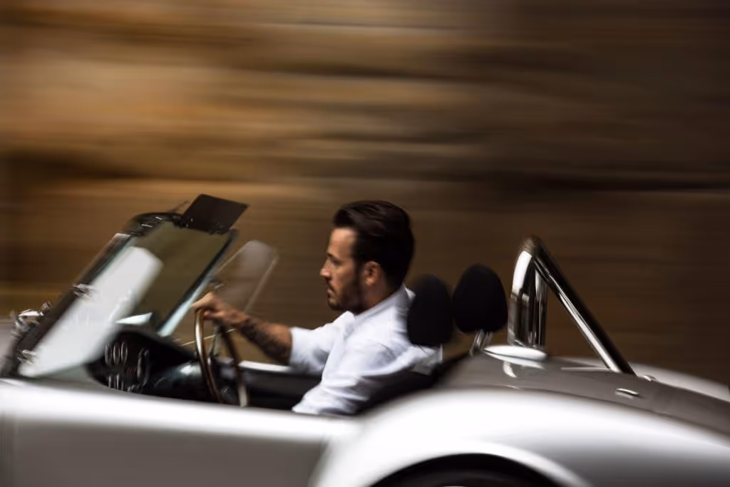 Man in white shirt driving a silver convertible car at speed with motion blur background.