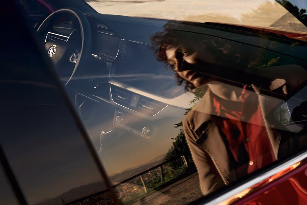 Reflection of a woman with curly hair in a red blouse and brown jacket looking into a car window, showing the car's interior and steering wheel.