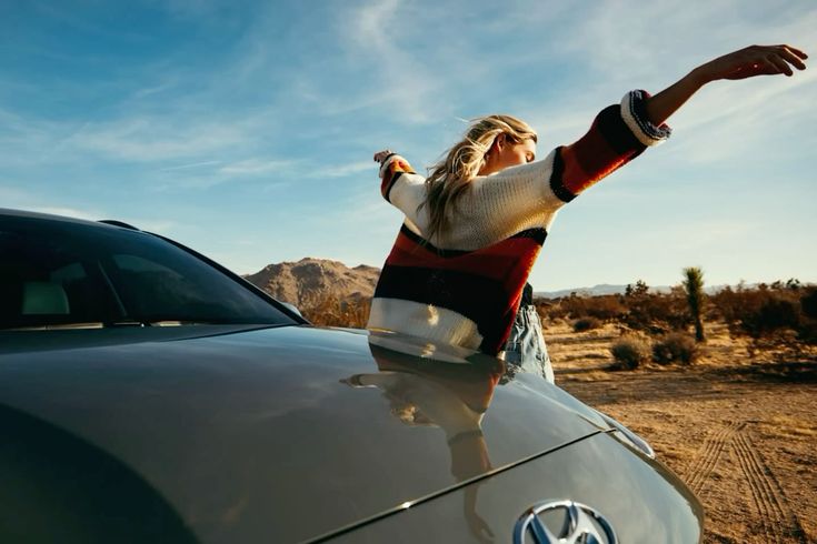 Person with arms outstretched beside a silver Volkswagen car in a desert landscape with mountains and blue sky.