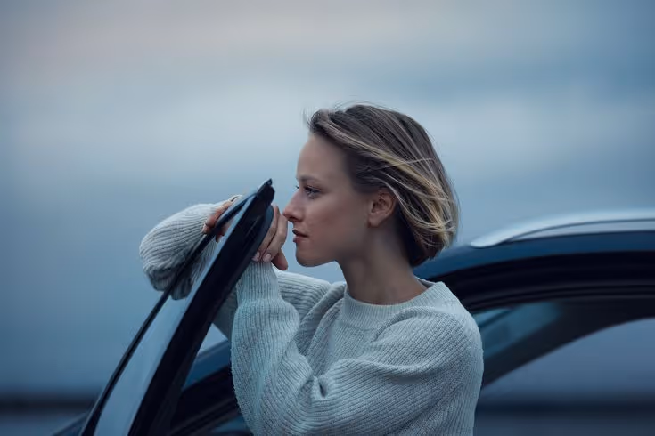 Woman in a gray sweater leaning on the open door of a car with a contemplative expression against a blurred outdoor background.