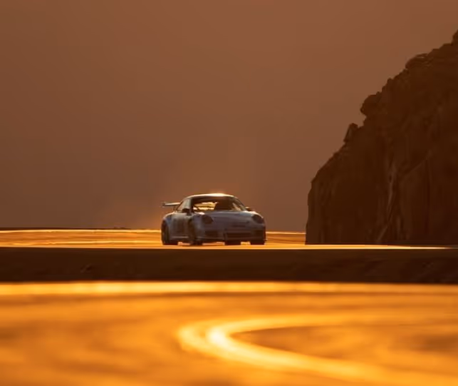 Blue sports car driving on a road at sunset with warm golden light reflecting on the surface and rocky cliffs on the right.