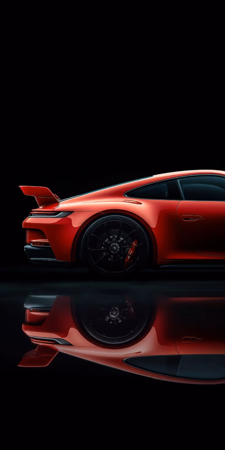 Side view of a red sports car with a raised rear spoiler and black wheels reflected on a glossy surface.