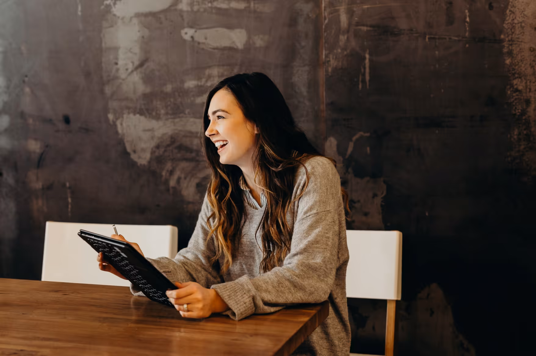 Smiling woman with long hair in a gray sweater holding a tablet and pen while sitting at a wooden table.