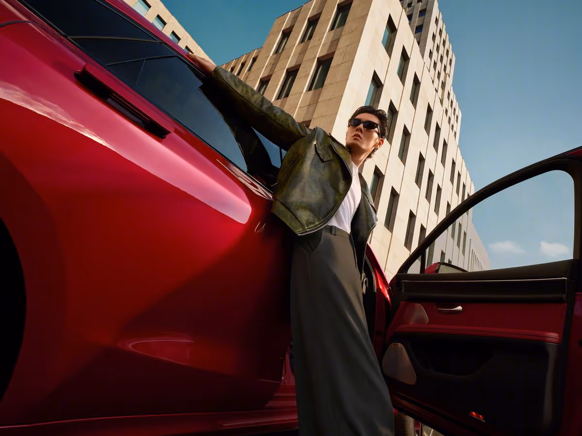 Fashionable person wearing sunglasses and a leather jacket leaning against an open red car door with a tall building in the background.