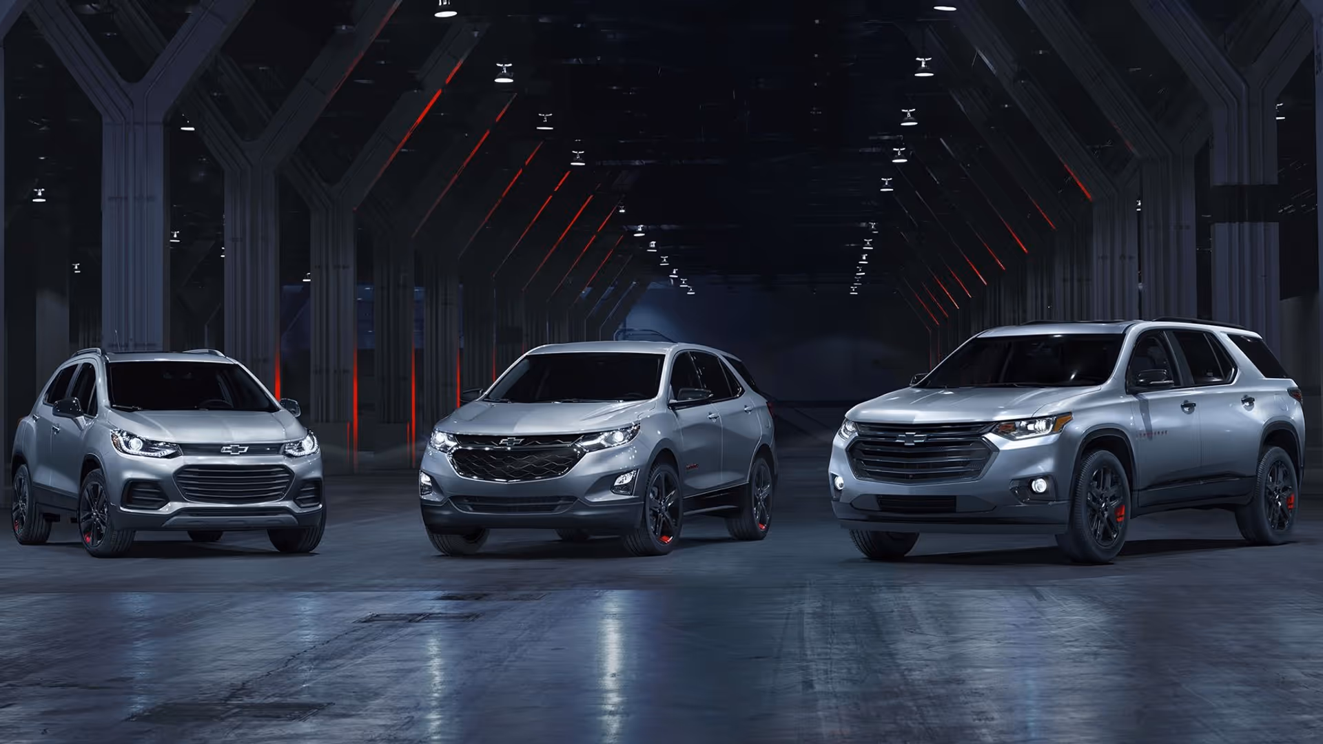 Three silver Chevrolet SUVs with black wheels and red accents parked inside a dark industrial building with red lighting accents.