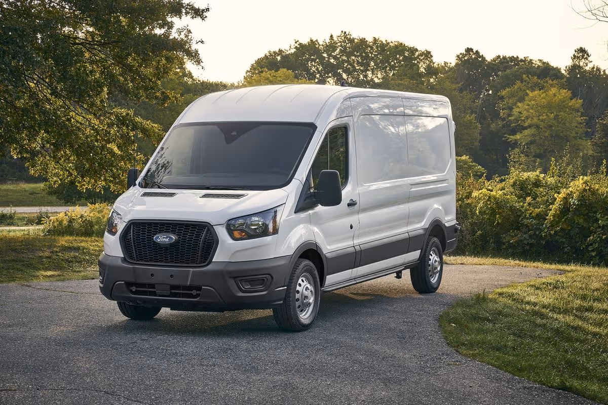 White Ford Transit van parked on a paved driveway surrounded by green trees and grass.
