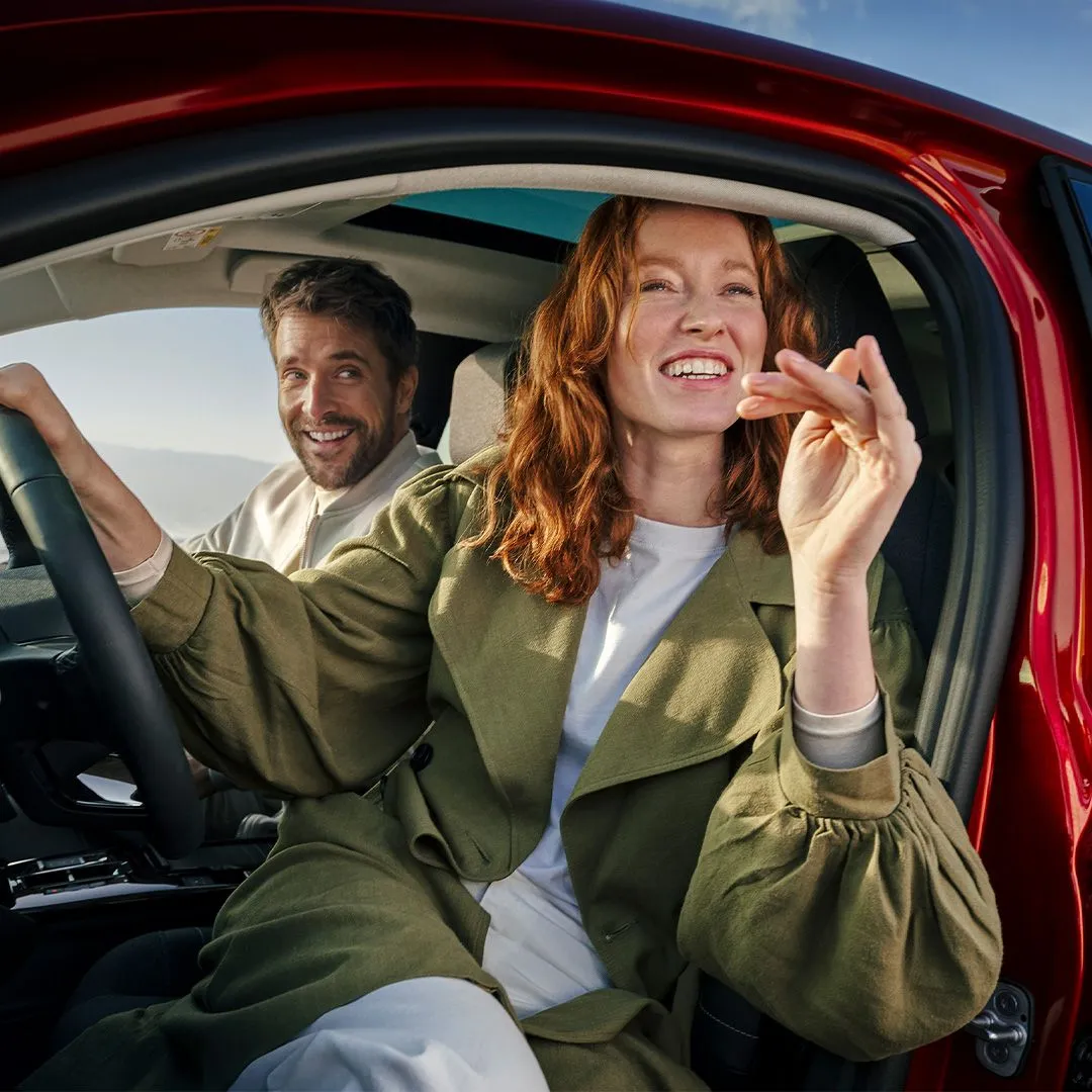 Smiling woman in green jacket sitting in the driver's seat of a red car with a smiling man in the passenger seat.
