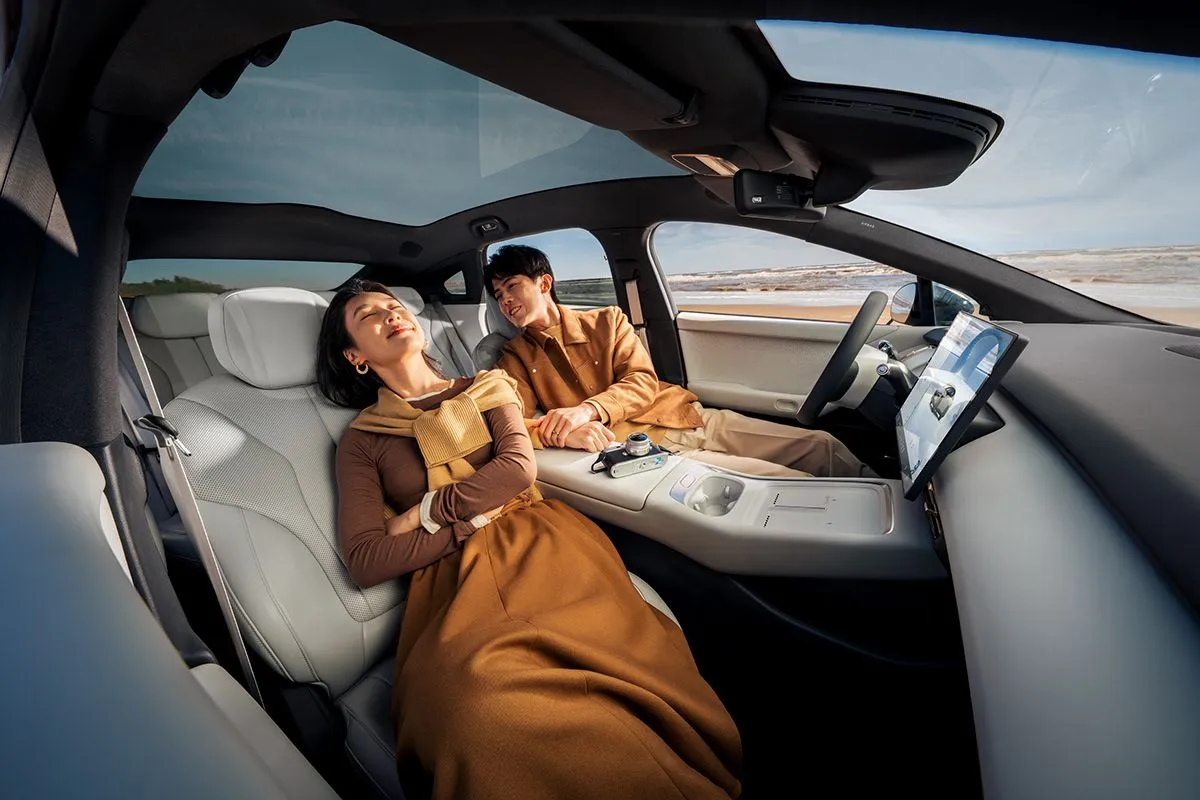 Two people relaxing inside a modern car with light-colored leather seats and a large touchscreen dashboard, near a beach.