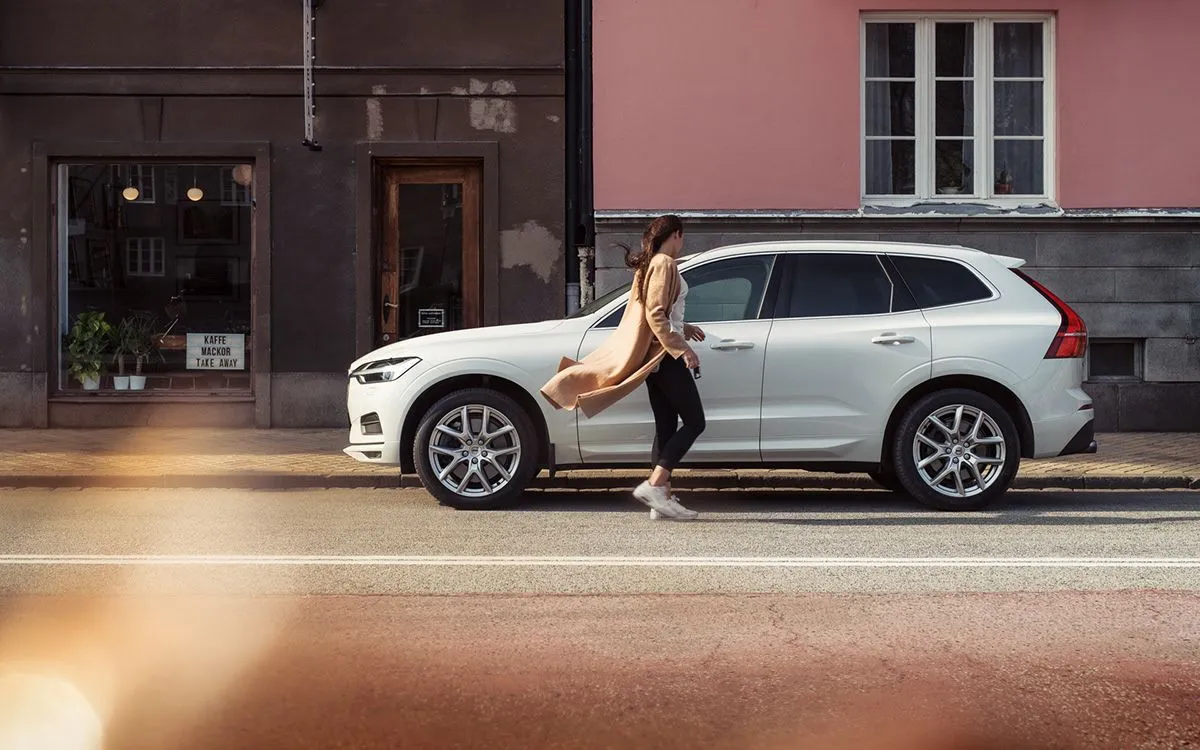 Woman in a beige coat walking past a white SUV parked on a city street in front of buildings with windows.