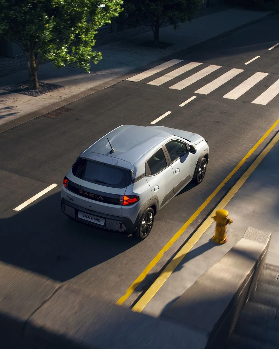 Silver Dacia Spring car driving on a city street near a crosswalk and a yellow fire hydrant.