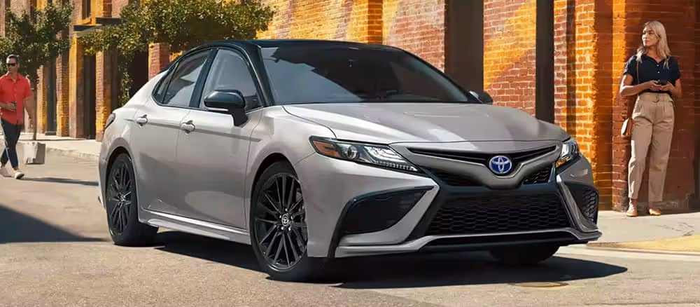 Silver Toyota Camry parked on a sunlit urban street with brick walls, with a man and woman standing on the sidewalk.