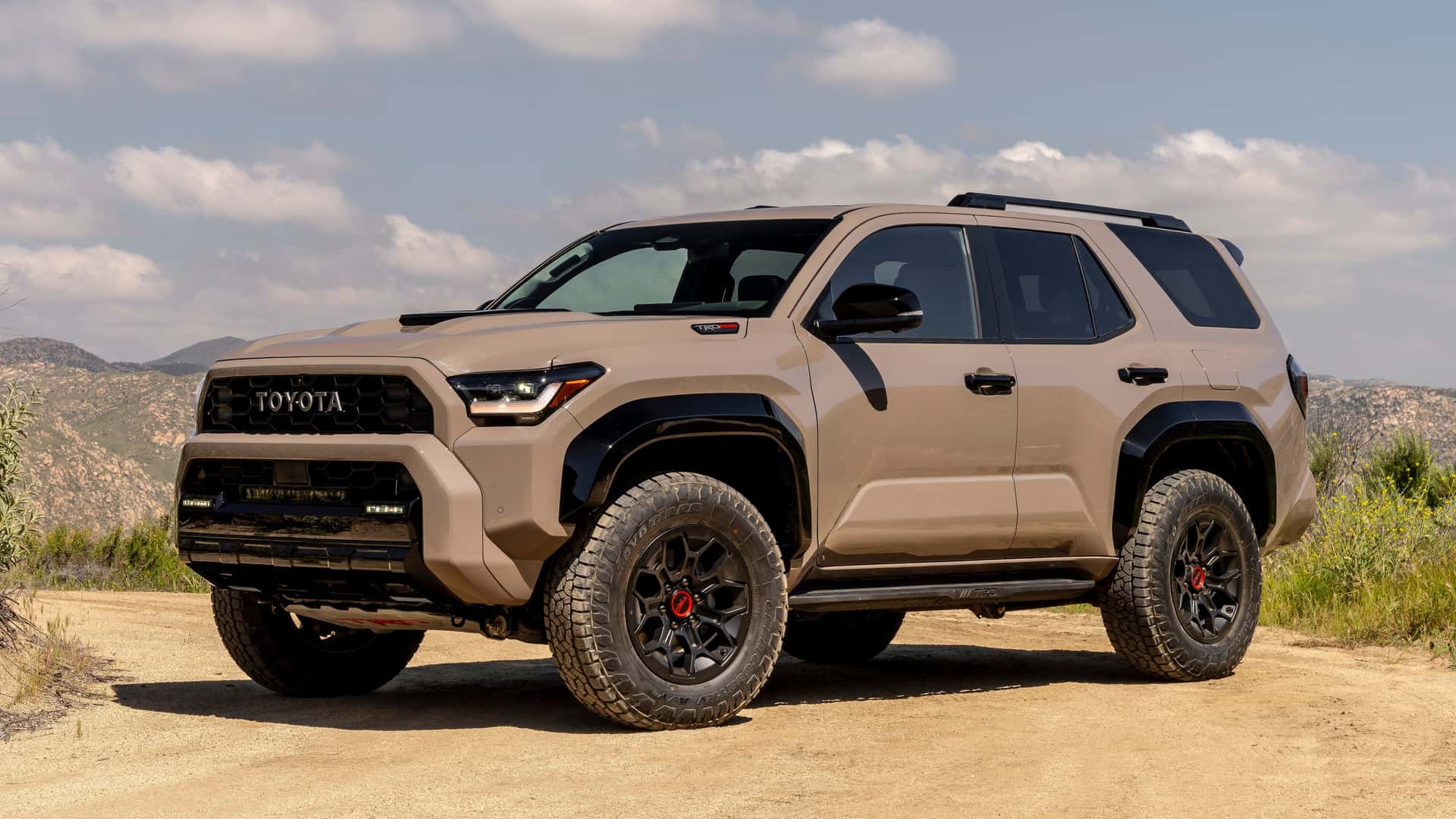 2025 Toyota 4Runner TRD Pro tan off-road SUV parked on dirt with mountains and cloudy sky in the background.