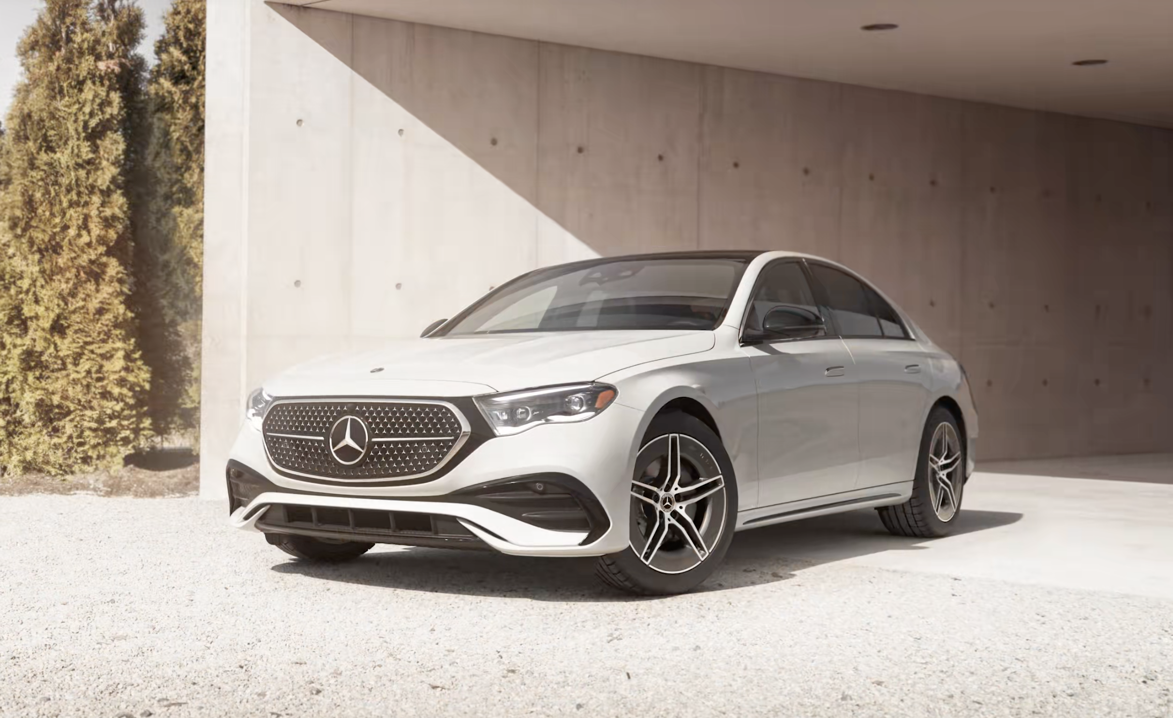 White Mercedes-Benz sedan parked on gravel near a modern concrete building with tall trees in the background.