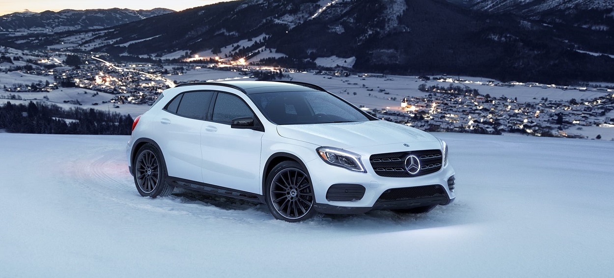 White Mercedes-Benz SUV parked on snow with a snowy village and mountains in the background at dusk.