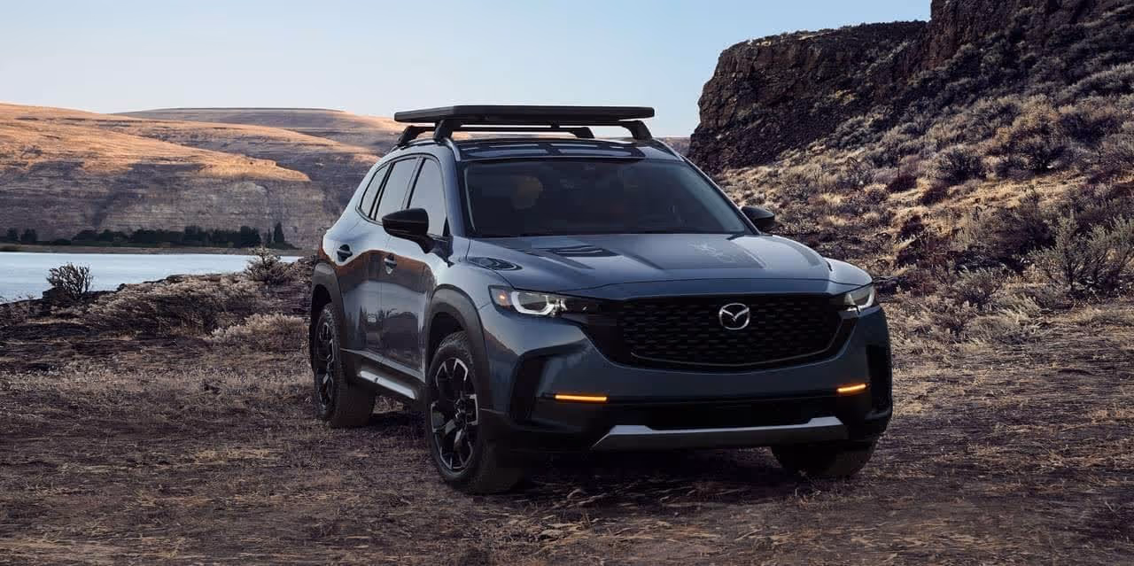 Dark gray Mazda CX-50 SUV parked on rugged terrain with hills and a river in the background.