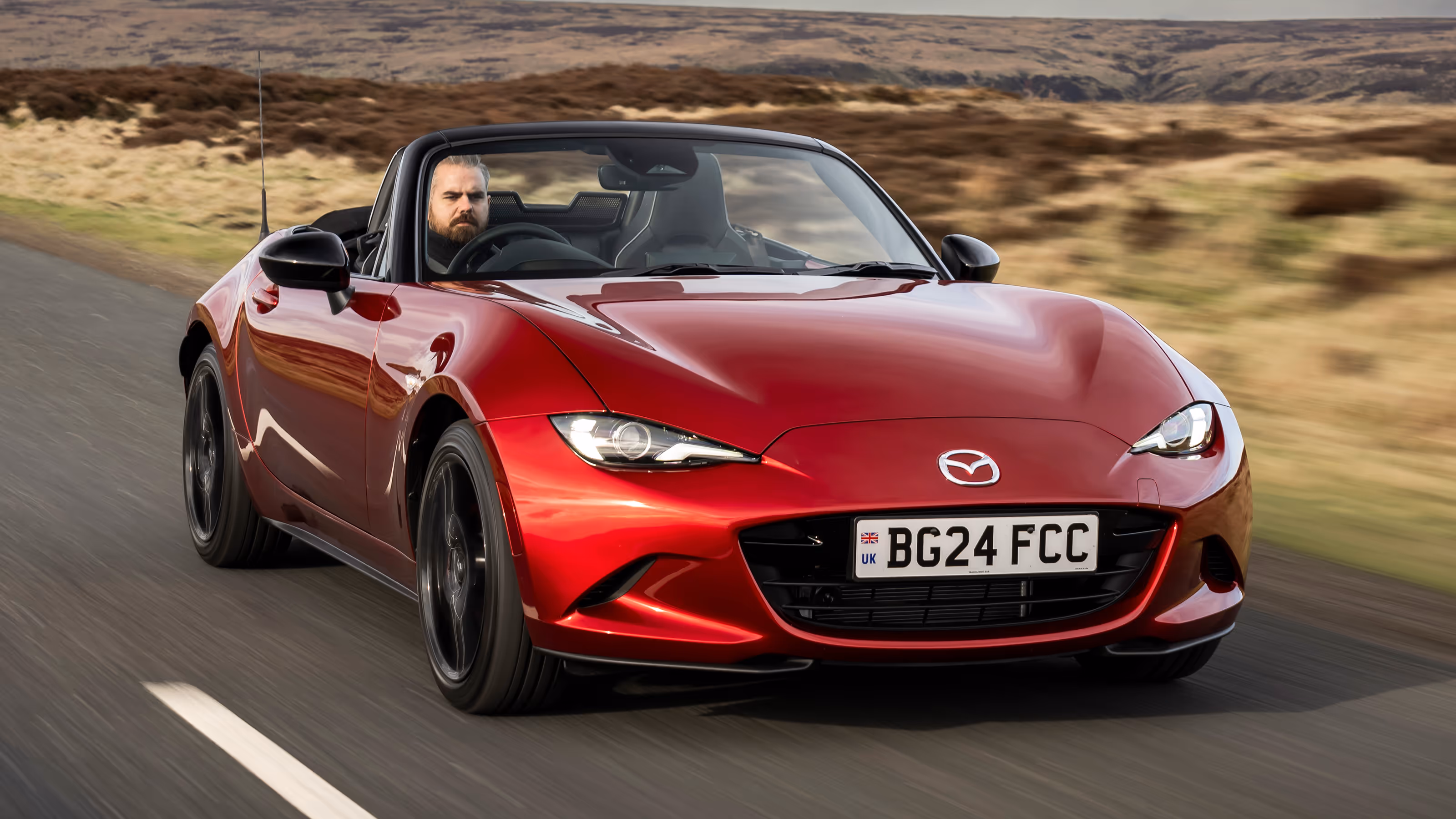 Red Mazda MX-5 convertible driving on a rural road with a man behind the wheel.